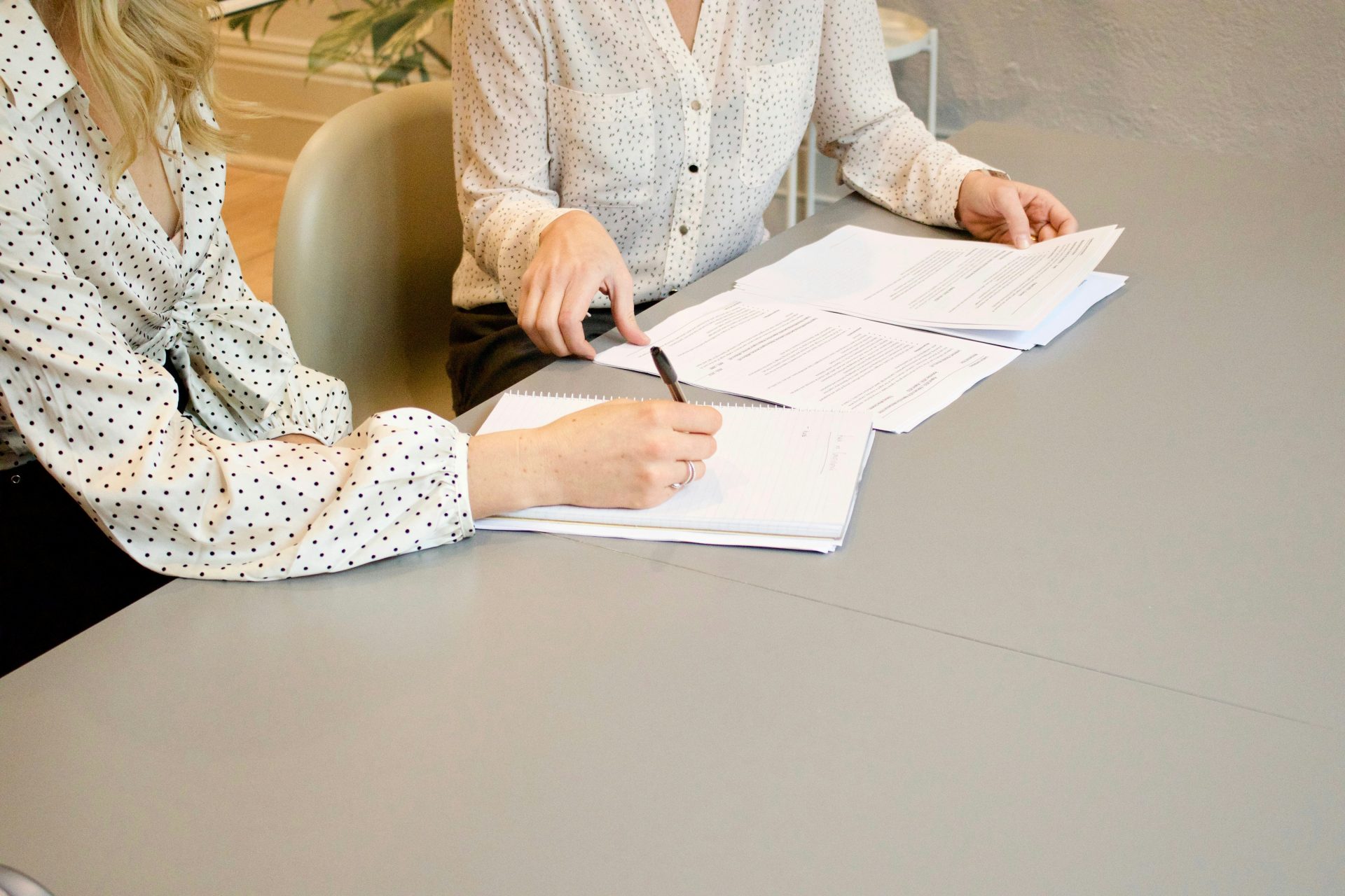 Mujer firmando en papel blanco de impresora junto a una mujer a punto de tocar los documentos